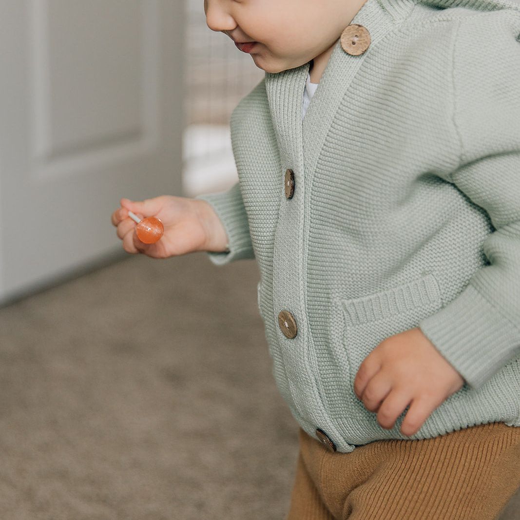 Child wearing a light green cardigan with buttons and ribbed brown leggings indoors
