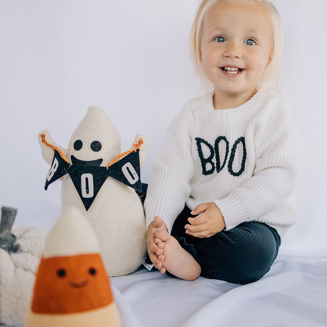 Little girl wearing a white organic cotton baby sweater with hand-stitched “BOO” lettering in black wearing black pants sitting in front of a while backdrop with halloween decor around her