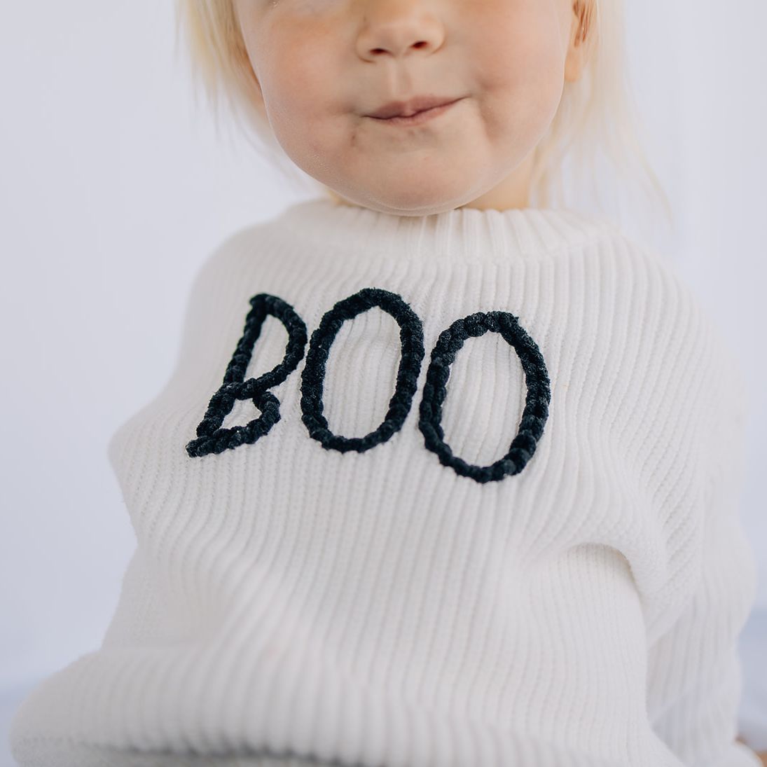 Close up of little girl wearing a white organic cotton baby sweater with hand-stitched “BOO” lettering in black in front of a while backdrop