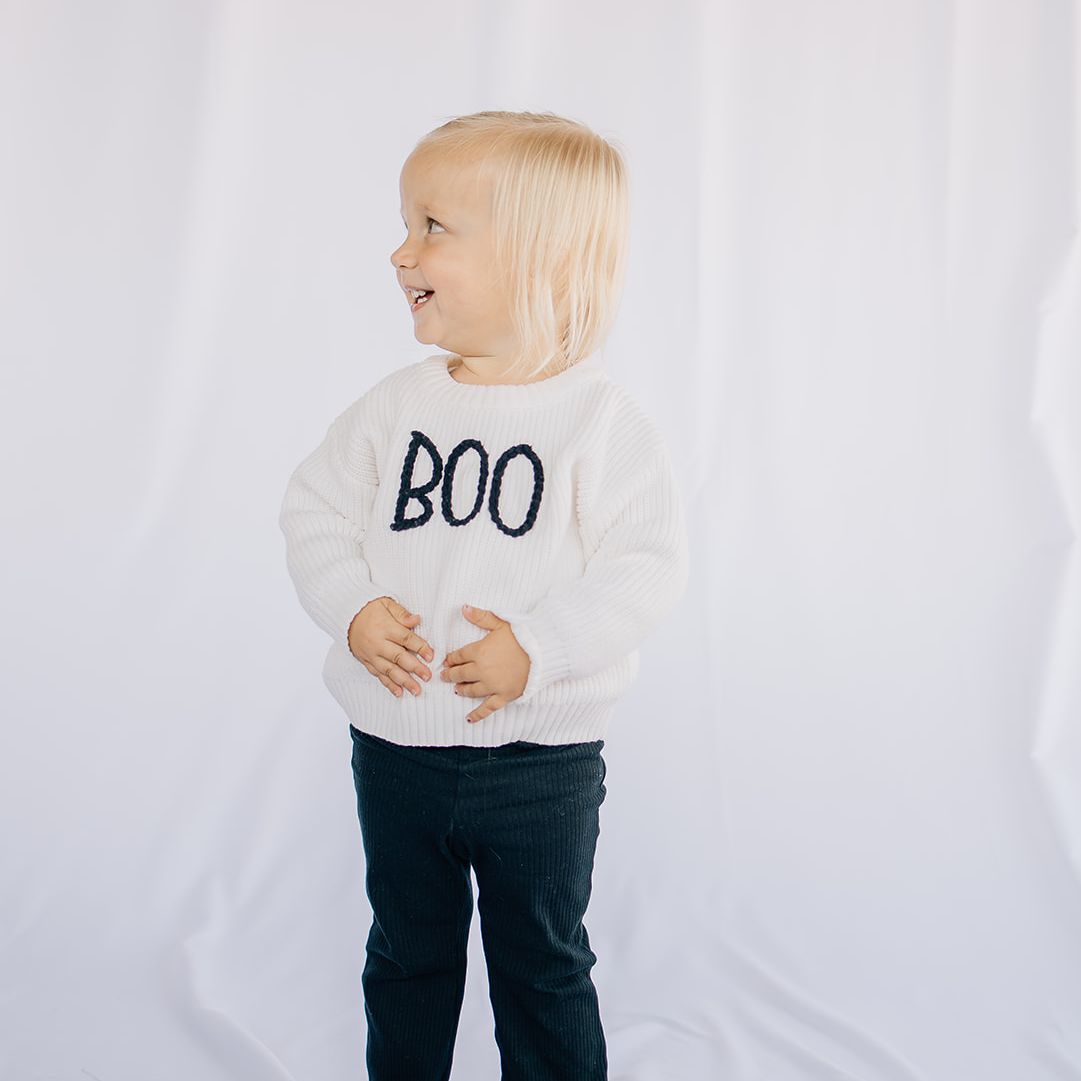 Little girl wearing a white organic cotton baby sweater with hand-stitched “BOO” lettering in black wearing black pants standing in front of a while backdrop
