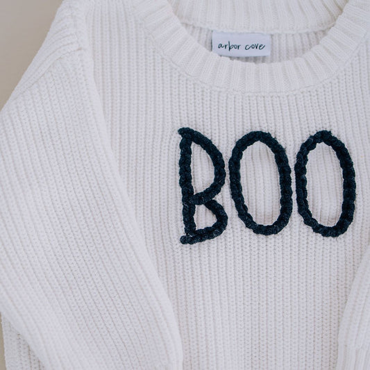 Close up of white organic cotton baby sweater with hand-stitched “BOO” lettering in black
