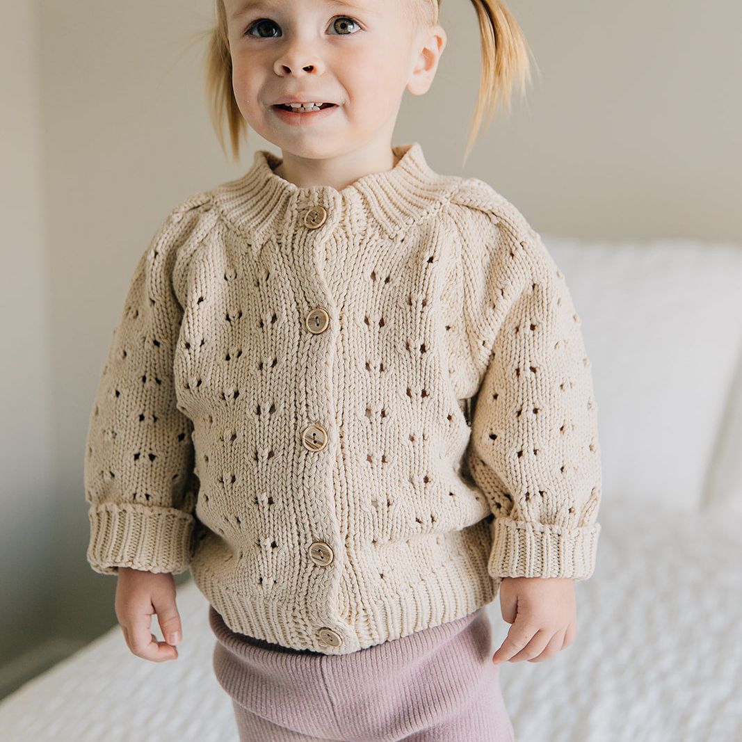 Child wearing a beige knitted cardigan and pink pants on a white background