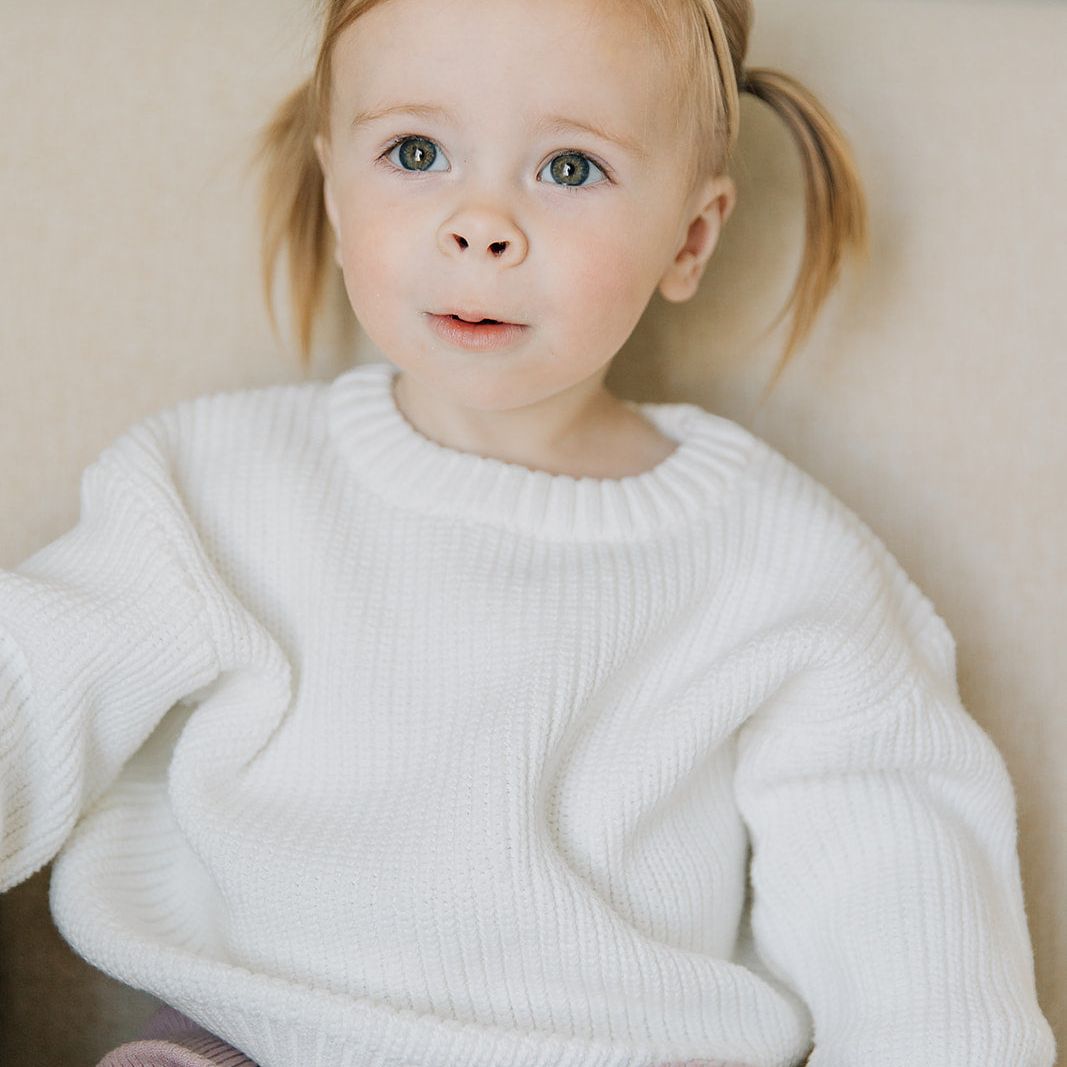 Young child wearing a white organic cotton sweater and pink pants, sitting on a beige couch.