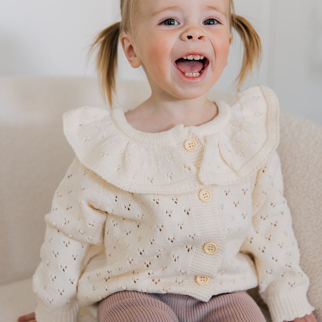 Child wearing a cream-colored knit pointelle cardigan sweater with buttons, sitting on a white couch.