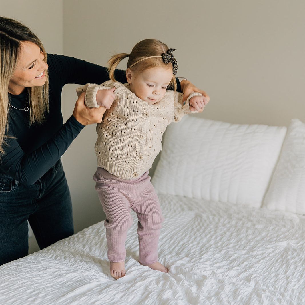 Woman helping a young child wearing a chunky knit sweater in oat color and mauve ribbed leggings stand on a bed