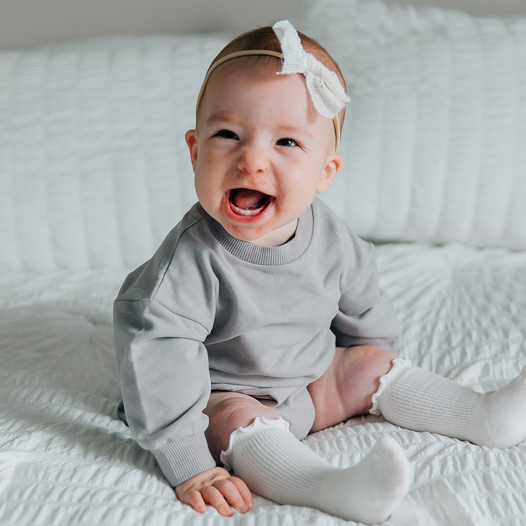 Baby wearing a gray long sleeved romper and white headband sitting on a white bed.