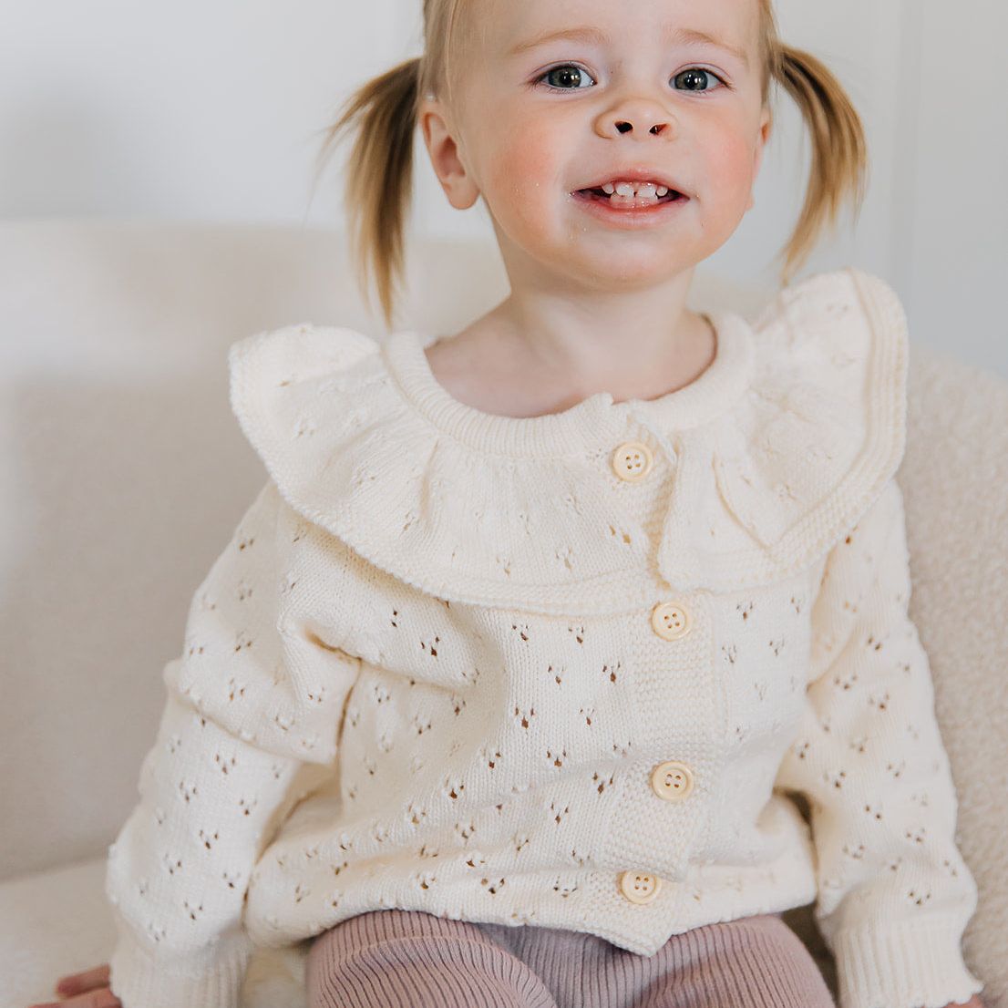 Young girl wearing a white pointelle cardigan with buttons and ruffled neck on a light background