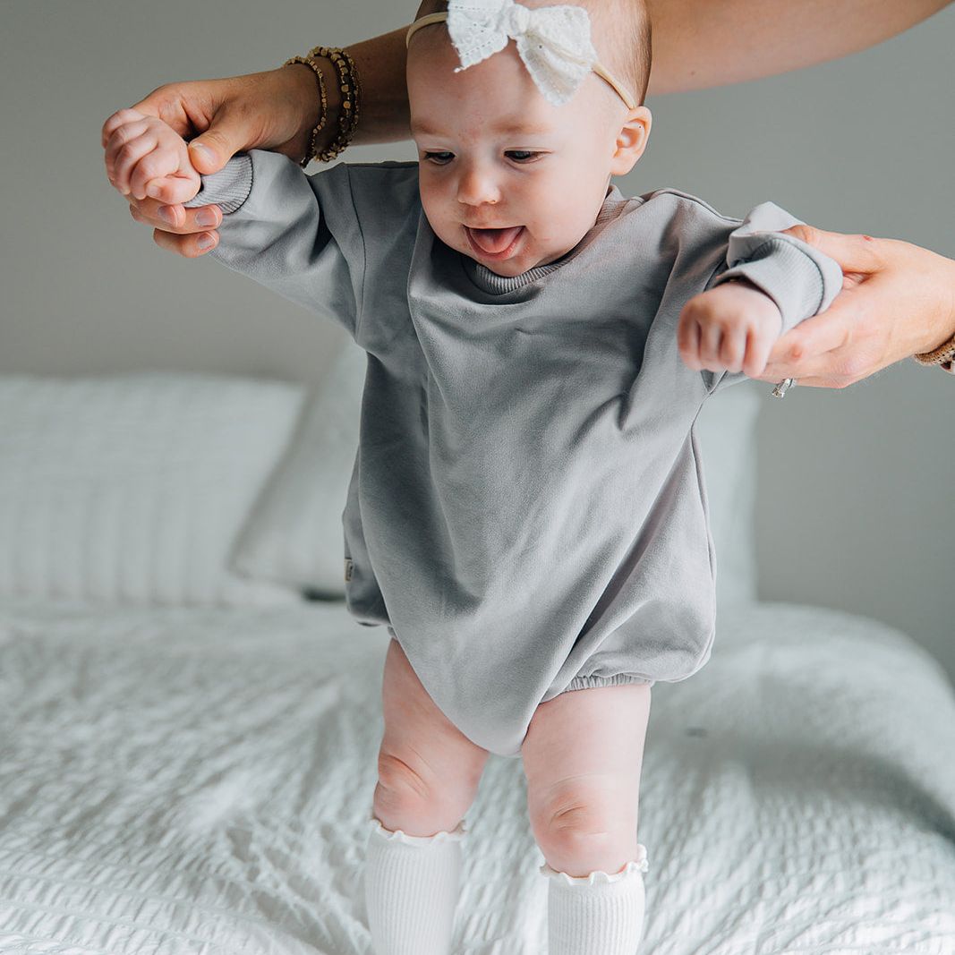 Baby in a gray long sleeved romper with a white bow standing on a bed