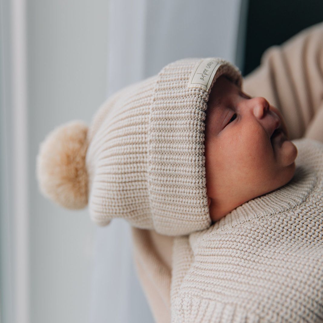 Baby wearing a oat colored knit outfit with a matching beanie hat with a pom pom indoors.