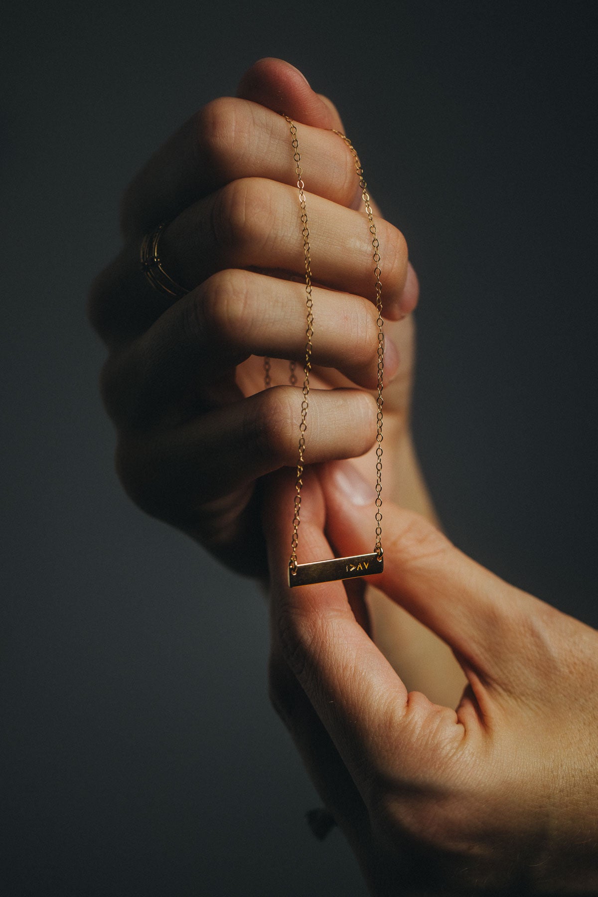 Hand holding a gold necklace with a bar with symbol I am greater than my highs and lows against a dark background