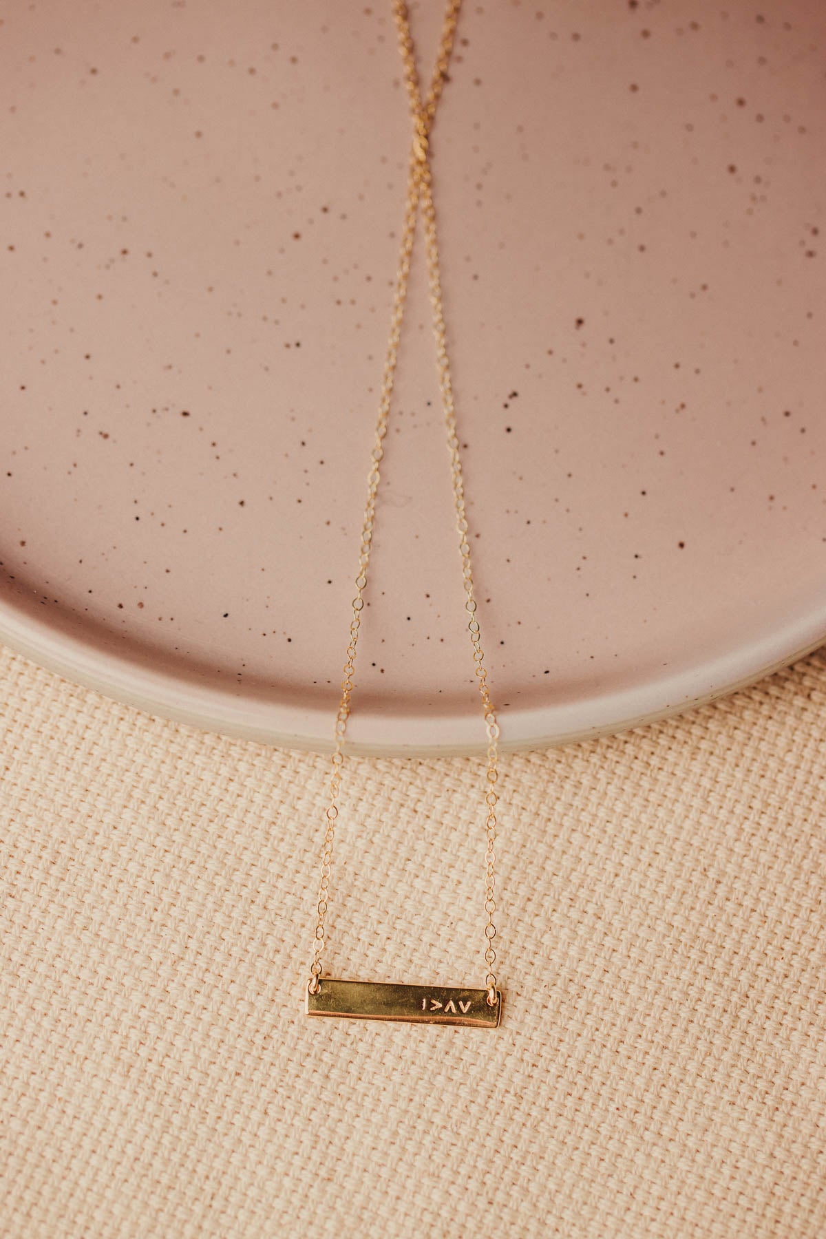Gold necklace with a bar pendant on a beige surface with symbol I am greater than my highs and lows