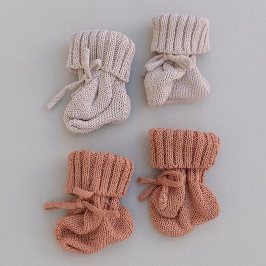 Two pairs of knitted baby booties in oat and rust on a light gray background