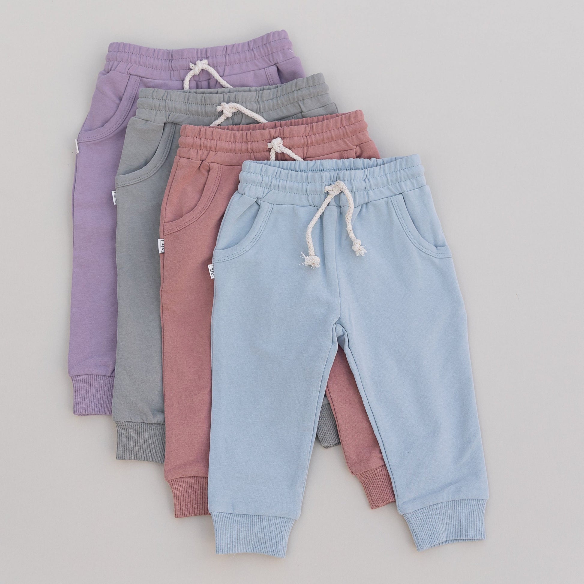 Four pairs of children's jogger pants in purple, green, pink and blue stacked on a light gray background.