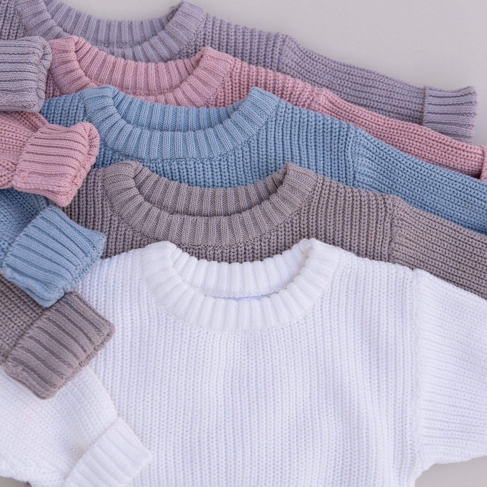 Stack of organic cotton baby sweaters in grey, pink, blue, brown and white on a neutral background