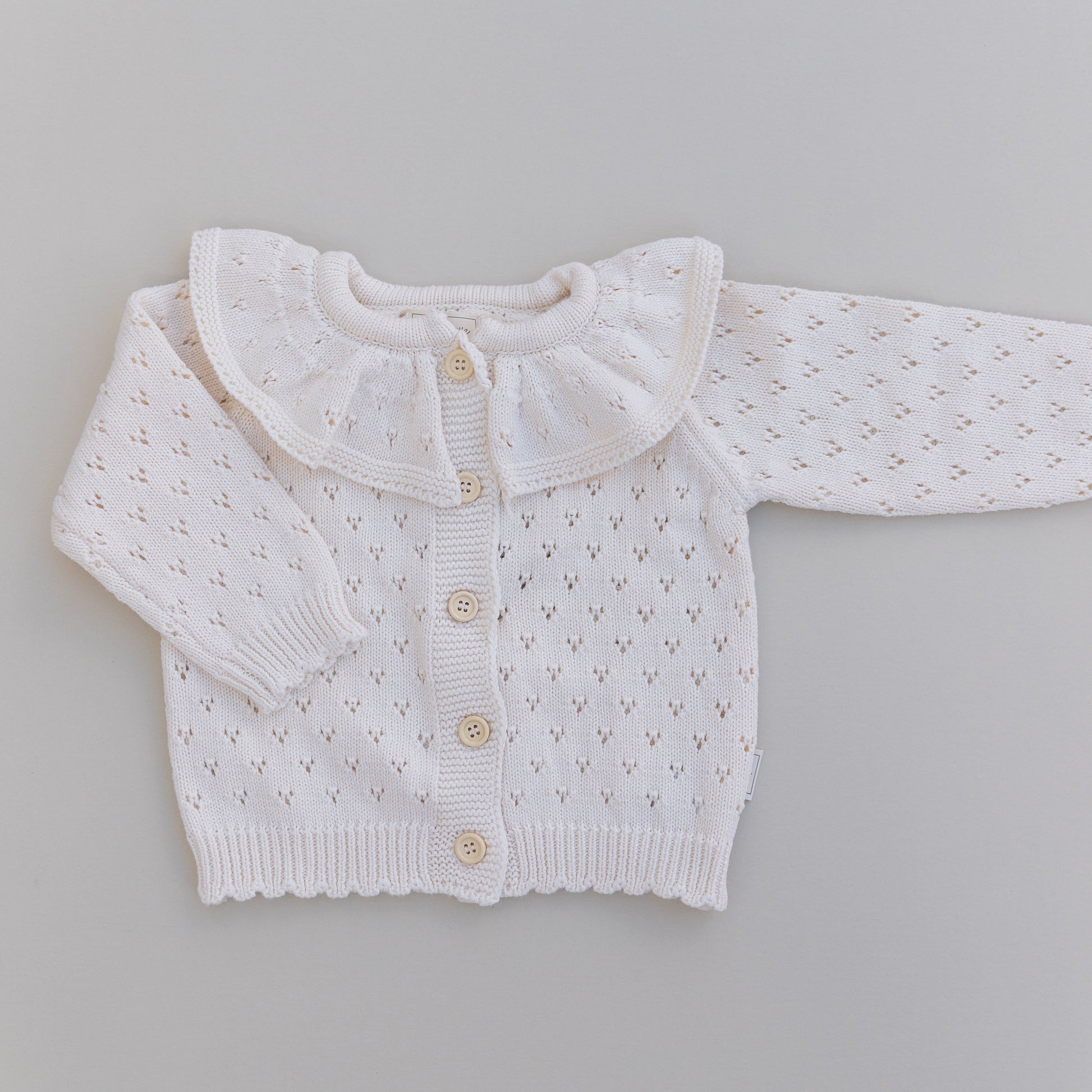 Cream pointelle baby cardigan with a ruffled collar on a gray background