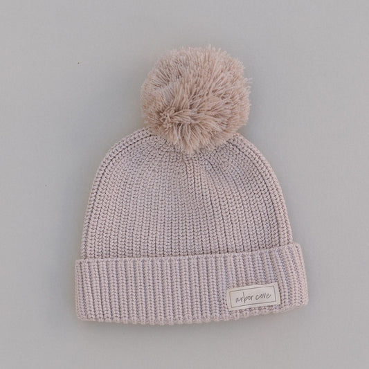 Oat colored knit baby beanie with pom-pom on a light gray background