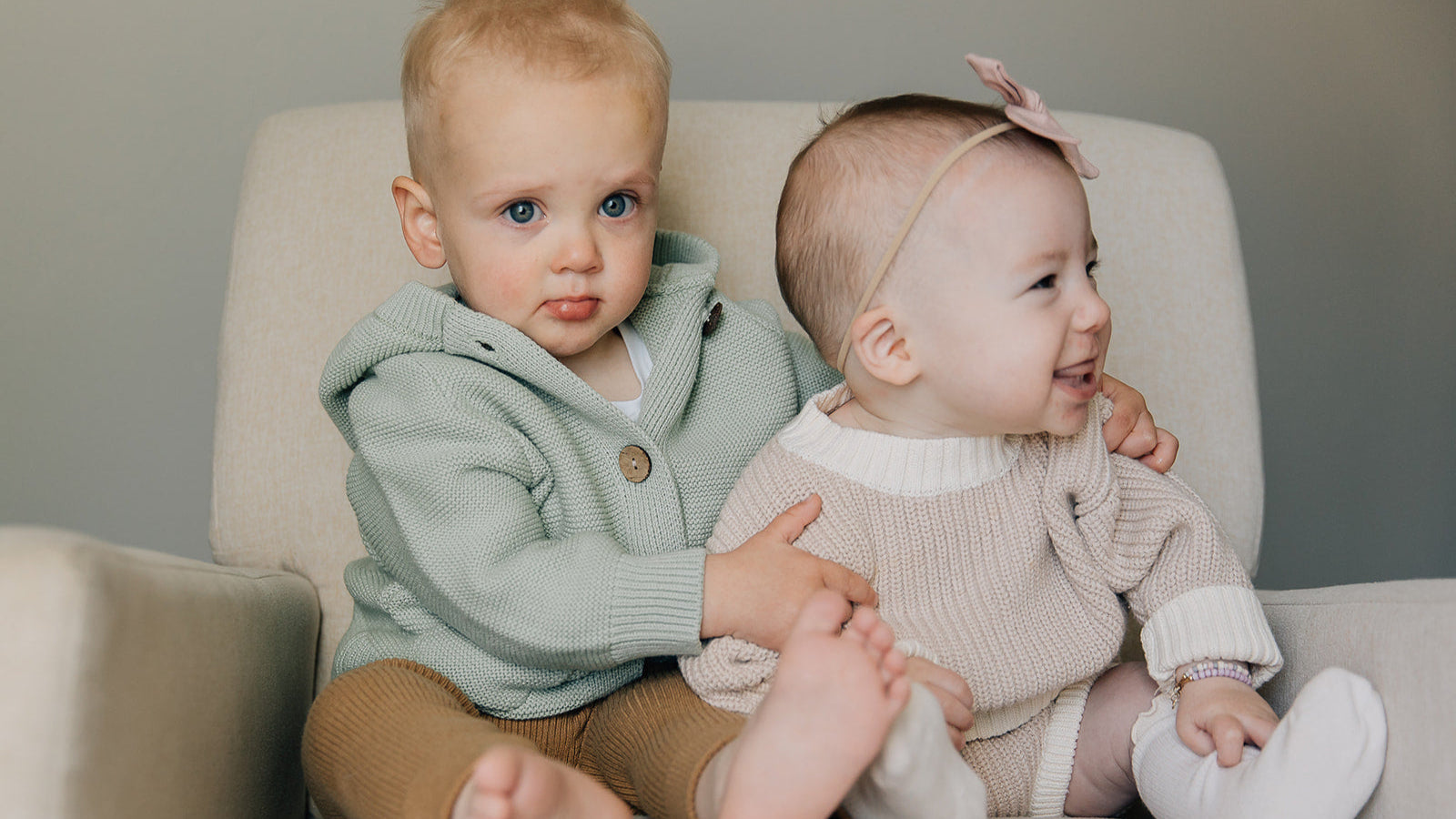 Two young children sitting on a couch, one wearing a green sweater and the other in a oat colored outfit with a headband.
