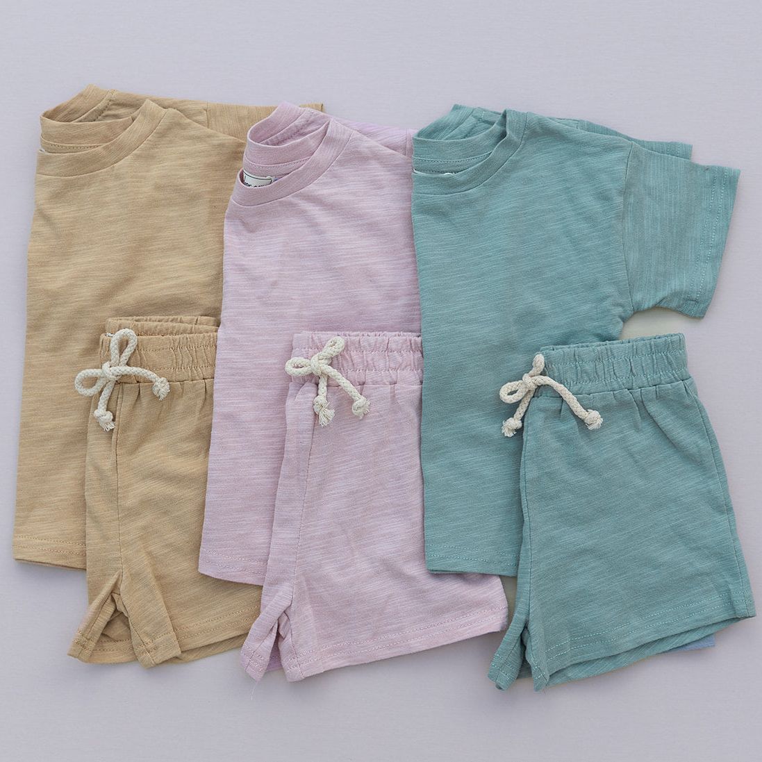 Three pairs of children's shorts in mustard, pink, green on a light gray background.